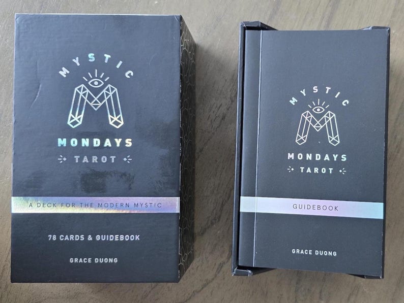 May include: Two black boxes of "Mystic Mondays Tarot" cards and guidebook. The boxes have a holographic logo and text that reads "A Deck for the Modern Mystic" and "78 Cards & Guidebook". The author's name, Grace Duong, is also printed on the boxes.