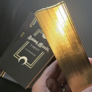 May include: A deck of tarot cards with gold gilded edges, held in hand, next to a black guidebook. The guidebook has the words "Anima Mundi Tarot" in gold lettering. The cards have a shiny, reflective surface.