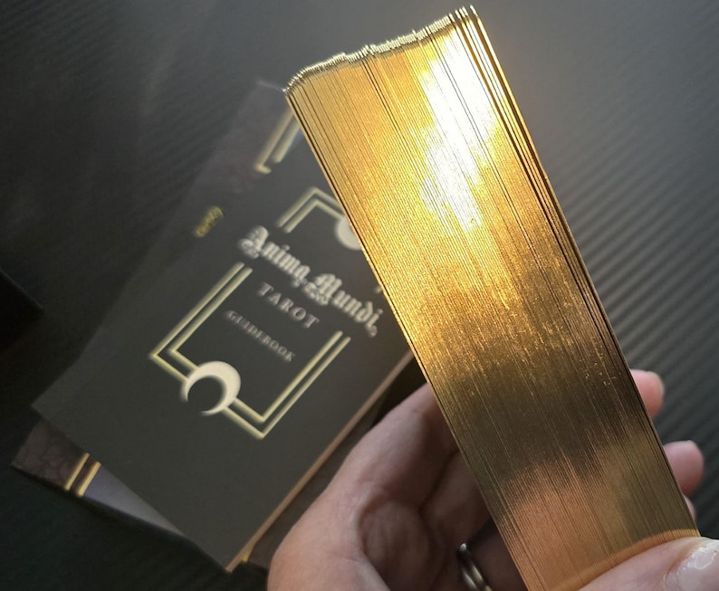 May include: A deck of tarot cards with gold gilded edges, held in hand, next to a black guidebook. The guidebook has the words "Anima Mundi Tarot" in gold lettering. The cards have a shiny, reflective surface.