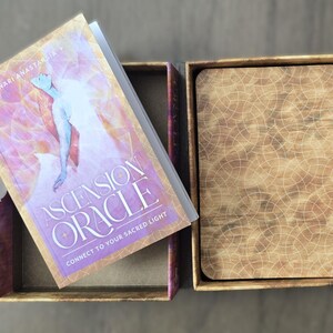 May include: A closed box with a wooden lid and a book titled "Ascension Oracle: Connect to Your Sacred Light" by "Nari Anastasia" on top of it.
