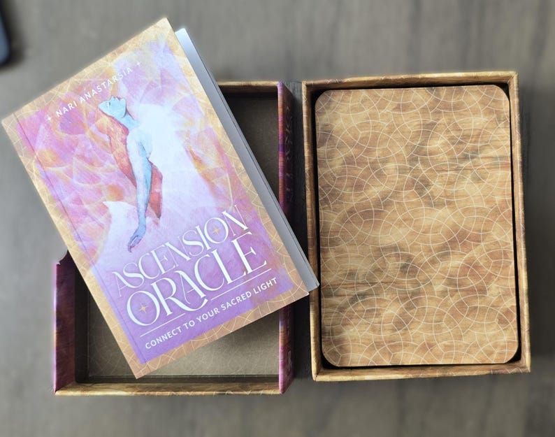 May include: A closed box with a wooden lid and a book titled "Ascension Oracle: Connect to Your Sacred Light" by "Nari Anastasia" on top of it.