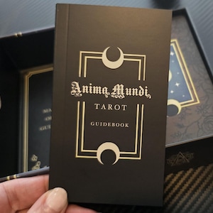May include: A black tarot guidebook with gold lettering and a crescent moon design. The book is titled "Anima Mundi, Tarot, Guidebook." The guidebook is in a black box with a floral design and a starry night illustration.