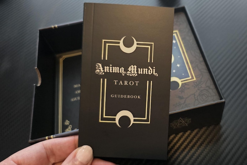 May include: A black tarot guidebook with gold lettering and a crescent moon design. The book is titled "Anima Mundi, Tarot, Guidebook." The guidebook is in a black box with a floral design and a starry night illustration.