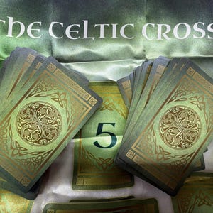 May include: A deck of cards with green and gold Celtic knot designs on the backs, arranged on a green fabric cloth featuring the text THE CELTIC CROSS and the number 5. The cards display intricate circular patterns and geometric borders.