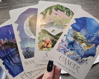 May include: A set of tarot cards with watercolor illustrations of astrological signs. The cards feature a blue and purple color scheme with gold accents. The cards are spread out in a fan shape, revealing the signs Pisces, Cancer, and Sagittarius.