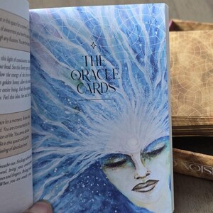 May include: A close-up of a blue and white oracle card deck with the title "The Oracle Cards" printed on the front. The card features a watercolor illustration of a woman with her eyes closed and a crown of blue and white branches.