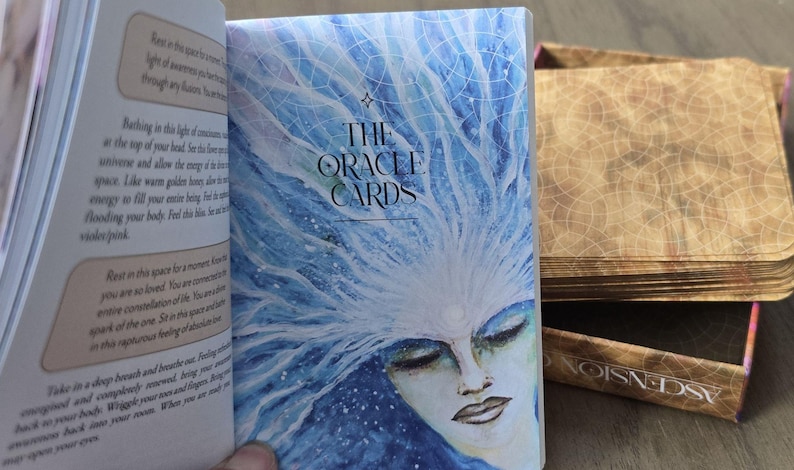 May include: A close-up of a blue and white oracle card deck with the title "The Oracle Cards" printed on the front. The card features a watercolor illustration of a woman with her eyes closed and a crown of blue and white branches.