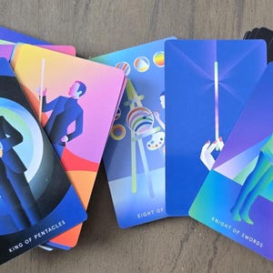 May include: A deck of tarot cards with colorful geometric illustrations. The cards feature figures and symbols, including the "King of Pentacles" and "Knight of Swords." The color palette includes blues, purples, and yellows. The cards are arranged on a wooden surface.
