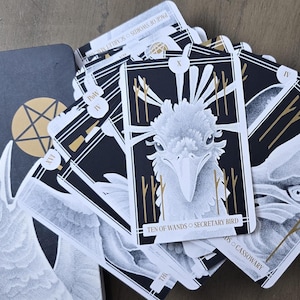 May include: The Avian Tarot deck and guidebook. The guidebook is dark gray with gold accents and the title "The Avian Tarot Guidebook". The cards are black and white with gold accents and bird illustrations. The cards are fanned out.