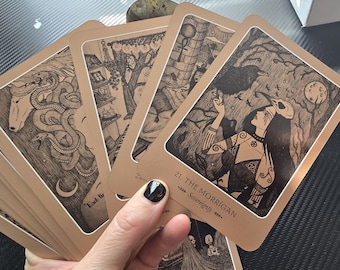 May include: A set of tarot cards with illustrations in black and white. The card in the foreground is titled "21. The Morrigan - Sovereignty". The card depicts a woman with a crow and a skull mask, wearing a long dress with intricate details. The cards have a brown background with a gold border.