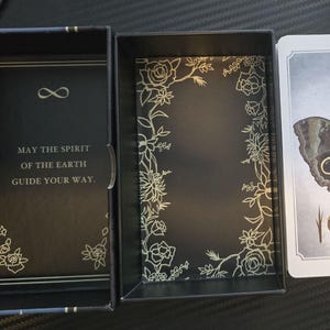 May include: A tarot card deck with a black box and cards. The box has gold floral designs and the text "MAY THE SPIRIT OF THE EARTH GUIDE YOUR WAY." The card features a butterfly and the words "The Magician."