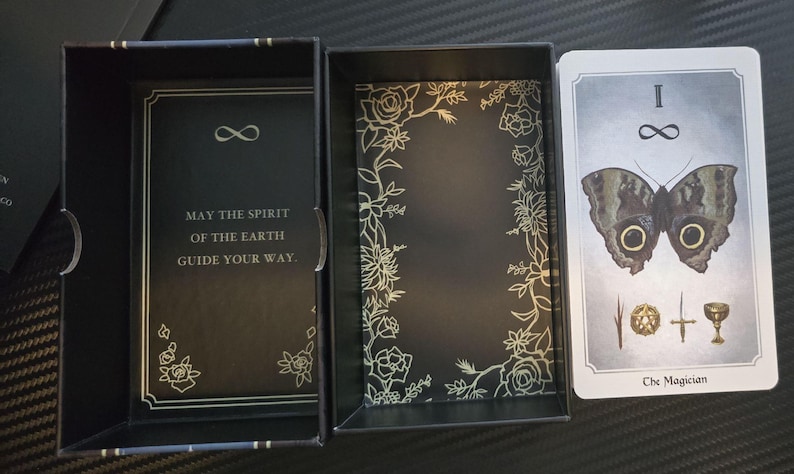 May include: A tarot card deck with a black box and cards. The box has gold floral designs and the text "MAY THE SPIRIT OF THE EARTH GUIDE YOUR WAY." The card features a butterfly and the words "The Magician."