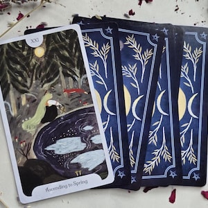 May include: A tarot card with a forest scene and the text "Ascending to Spring" is displayed with several other cards. The card's back has a navy blue background with crescent moons, stars, and botanical illustrations. The main card features a figure with long green hair.