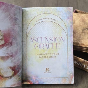 May include: A closed book with a watercolor design on the cover. The book is titled "Ascension Oracle" by "Nari Anastarsia". The text on the cover reads "Connect to your Sacred Light".