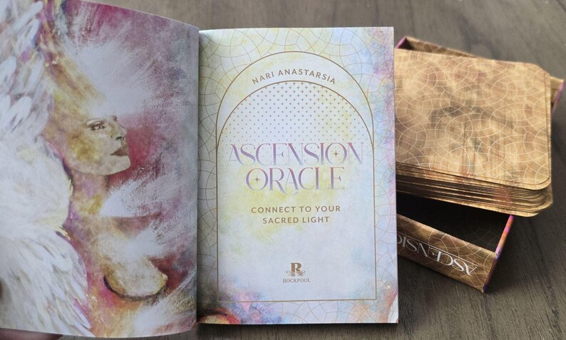 May include: A closed book with a watercolor design on the cover. The book is titled "Ascension Oracle" by "Nari Anastarsia". The text on the cover reads "Connect to your Sacred Light".