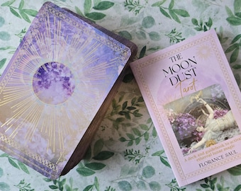 May include: A tarot deck and guidebook set. The deck features a purple and gold design with a celestial theme. The guidebook is light pink with the title "The Moon Dust Tarot" and an illustration of a woman.
