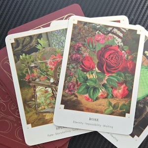 May include: A deck of tarot cards with burgundy backs and gold accents. Three cards are face up, featuring illustrations of a spinning wheel, a rose, and a green bottle. The card labeled "Rose" has the words "Eternity, Impossibility, Waiting."