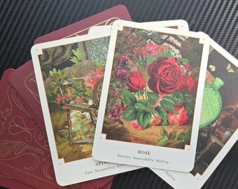 May include: A deck of tarot cards with burgundy backs and gold accents. Three cards are face up, featuring illustrations of a spinning wheel, a rose, and a green bottle. The card labeled "Rose" has the words "Eternity, Impossibility, Waiting."