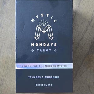 May include: A black tarot card deck titled "Mystic Mondays Tarot." The box features white geometric designs and the text "A Deck for the Modern Mystic." It includes 78 cards and a guidebook, authored by Grace Duong.