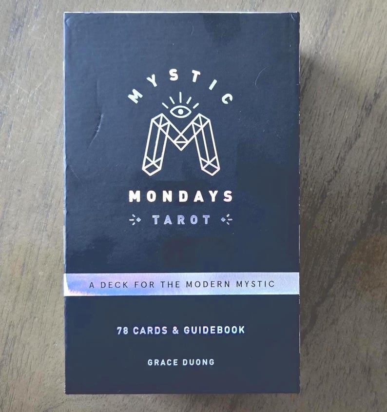 May include: A black tarot card deck titled "Mystic Mondays Tarot." The box features white geometric designs and the text "A Deck for the Modern Mystic." It includes 78 cards and a guidebook, authored by Grace Duong.