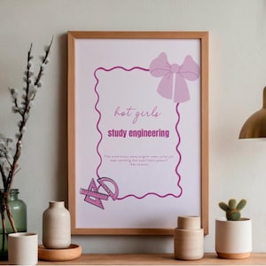 May include: A framed print with the text "hot girls study engineering" in a pink font, surrounded by a pink decorative border. The print includes a pink bow and engineering tools. The frame is a light brown color.