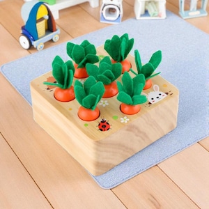 Peut inclure: Jeu en bois avec des carottes oranges et des feuilles vertes en feutre dans une boîte carrée en bois. La boîte est décorée d'une coccinelle, de fleurs et d'un lapin. Un jouet amusant et éducatif pour enfants.