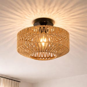 May include: A circular ceiling light with a woven rope shade. The light fitting has a black base and a natural tan rope shade. The light bulb is visible and lit, creating a warm ambience. The fixture is mounted on a white ceiling.
