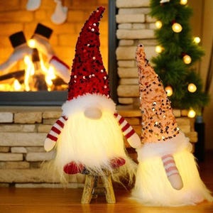 May include: Two illuminated Christmas gnomes. One gnome has a red sequined hat, white beard, and red and white striped arms and legs. The other gnome has a peach sequined hat and white beard. A Christmas tree and fireplace are in the background.