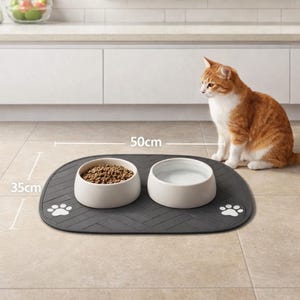 Könnte beinhalten: Eine graue Futtermatte für Haustiere mit zwei weißen Keramikschalen, eine gefüllt mit Trockenfutter und die andere mit Wasser. Die Matte ist 50 cm lang und 35 cm breit, mit Pfotenabdruckdetails. Eine rot-weiße Katze sitzt in der Nähe.