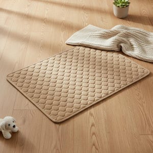 May include: A beige rectangular pet bed with a quilted, honeycomb pattern. A light-coloured blanket and a small potted succulent are visible in the background. A white stuffed dog toy is in the lower left corner. The bed is on a wooden floor.