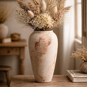May include: A tall, textured vase with a dried floral arrangement. The vase is off-white with brown accents. The arrangement includes wheat, fluffy beige elements, and small white flowers. The vase sits on a wooden table.