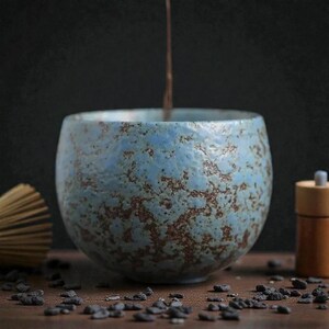 Peut inclure: Un bol à thé en céramique bleu avec une surface texturée et des accents bruns. Le bol est rond et repose sur une surface en bois. Un liquide coule dans le bol. Accessoires de thé en arrière-plan.