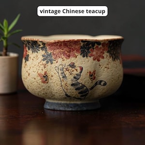 May include: Vintage Chinese teacup with a beige, textured finish. The rim features floral patterns in red, pink, and blue. A playful cat illustration is on the side. The teacup has a small, blue base.
