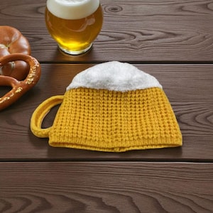 Può includere: Un cappello a forma di boccale di birra realizzato in tessuto giallo a maglia con una parte superiore bianca e soffice, che ricorda la schiuma della birra. Il cappello ha un manico giallo ed è esposto su una superficie di legno.