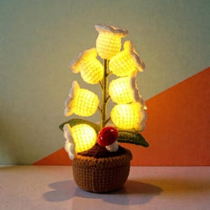Peut inclure: Une lampe fleur en crochet faite à la main avec des lumières jaunes en forme de fleurs. La lampe comprend un pot en crochet marron, des feuilles vertes et un petit champignon rouge. Le fond est un mélange de vert et d'orange.