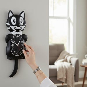 May include: A black and white cat-shaped wall clock with a white bow tie. The clock has a black face with white numbers and hands. The clock is hanging on a gray wall. The clock measures approximately 12 inches in height.