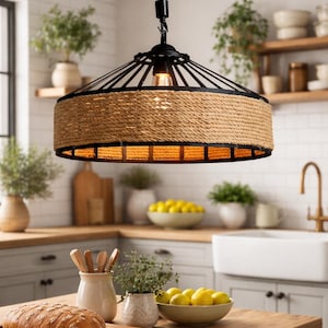 May include: A rustic pendant light with a black metal frame and a cylindrical shade wrapped in natural rope. The light hangs from a thick rope and illuminates a kitchen setting with wooden worktops and white cabinets.