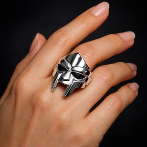 Peut inclure: Une bague argentée représentant un masque avec des orbites noires et un sommet pointu. La bague a une finition polie et est portée sur un doigt sur fond sombre. Le design du masque a une forme angulaire distinctive.