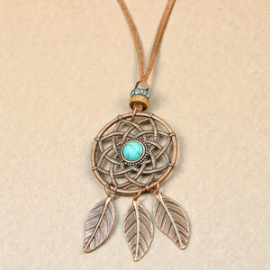 Peut inclure: Un collier pendentif attrape-rêves de couleur bronze avec une pierre turquoise au centre et trois breloques en forme de feuilles. Le collier a un cordon en cuir marron et un accent de perles décoratives.