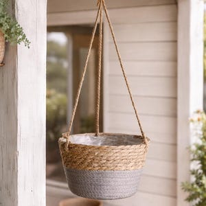 Peut inclure: Panier suspendu en osier avec une partie inférieure grise et une partie supérieure naturelle, suspendu par une corde. Le panier est conçu pour les plantes et est suspendu à un crochet en métal. L'arrière-plan comprend une structure en bois blanc et de la verdure.