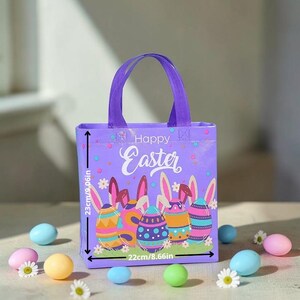 Peut inclure: Un sac fourre-tout violet sur le thème de Pâques avec l'inscription "Happy Easter" et des œufs de Pâques colorés. Le sac mesure 23 cm de haut et 22 cm de large. Des œufs pastel et des marguerites entourent le sac.