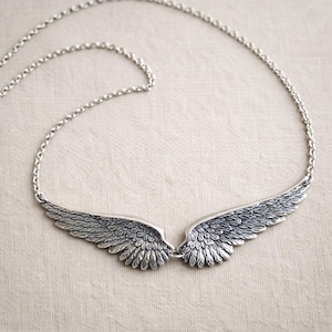 Peut inclure: Un collier argenté orné d'une paire de pendentifs en forme d'ailes détaillées. Les ailes sont finement conçues avec des détails de plumes et reliées au centre. Le collier a une chaîne délicate et est présenté sur un fond neutre.