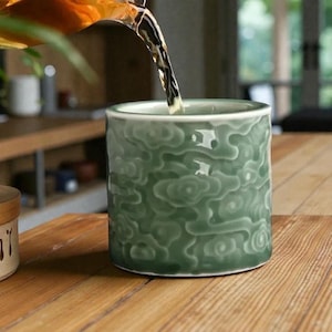 May include: A green ceramic tea cup with a cloud-like pattern is being filled with tea. The cup sits on a wooden table. The tea is a golden brown color. The cup is cylindrical and has a smooth, glossy finish.