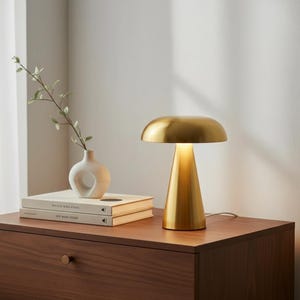 Peut inclure: Une lampe de table dorée avec un abat-jour arrondi et une base conique est éclairée sur une surface en bois. Un petit vase blanc avec une brindille de verdure et deux livres sont également sur la surface.