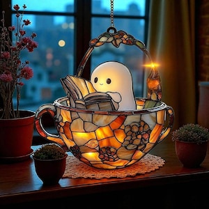 May include: A stained glass teacup lamp with a handle and a hanging decorative top. Inside the cup, a cartoon ghost figure reads a book. The lamp is lit, casting a warm glow. The teacup is decorated with amber and brown glass.