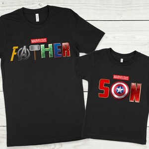 May include: Two black t-shirts with a Marvel theme. The larger shirt says "MARVELOUS FATHER" with the letters replaced by Marvel character symbols. The smaller shirt says "MARVELOUS SON" with the letters replaced by Marvel character symbols.