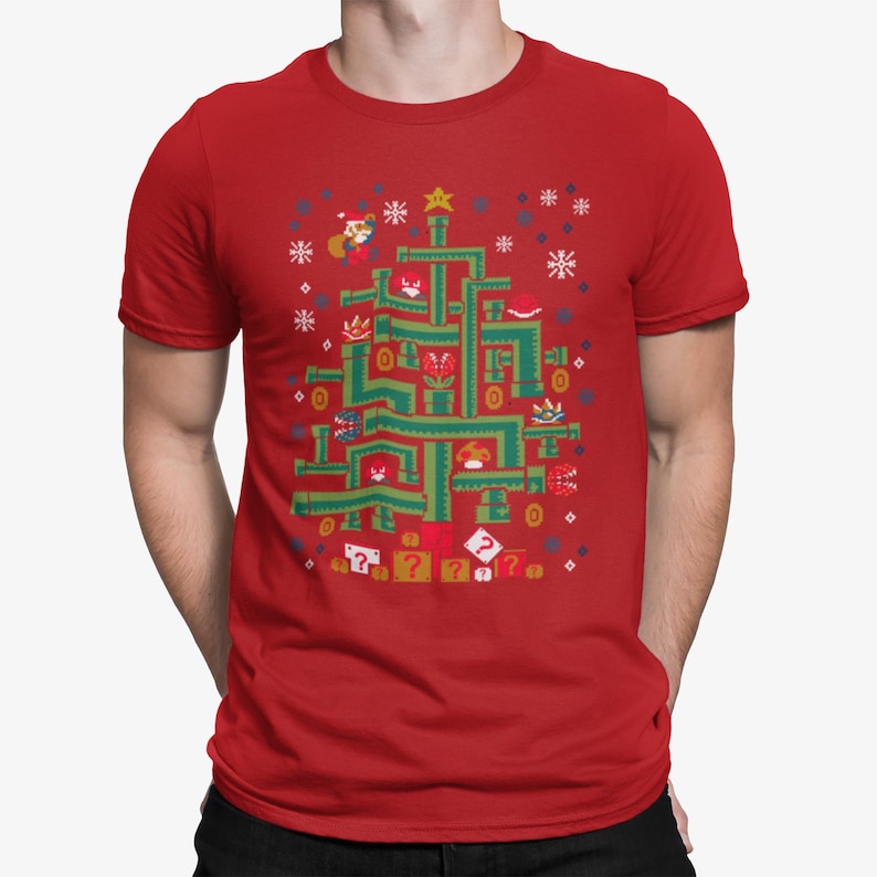 May include: Red t-shirt featuring a green Christmas tree design made of video game elements. The design includes characters, coins, and question mark blocks. Snowflakes and a star complete the festive look.