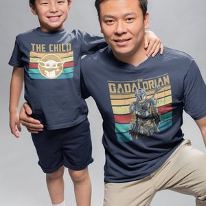 May include: A father and son wearing matching navy blue t-shirts with a vintage style graphic design. The son's shirt says "The Child" with a cartoon image of Baby Yoda. The father's shirt says "The  Dadalorian" with a cartoon image of a Mandalorian.