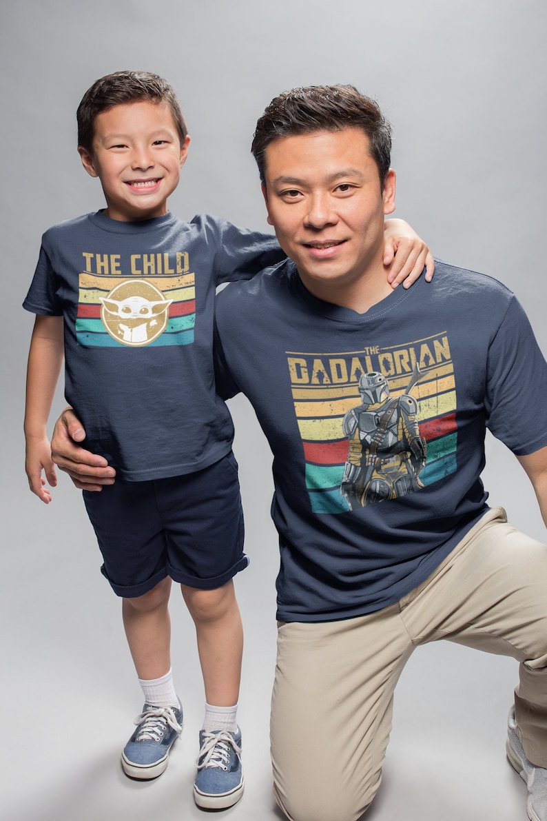 May include: A father and son wearing matching navy blue t-shirts with a vintage style graphic design. The son's shirt says "The Child" with a cartoon image of Baby Yoda. The father's shirt says "The  Dadalorian" with a cartoon image of a Mandalorian.