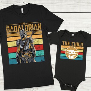 May include: Two black t-shirts with a retro design. The larger shirt features a Mandalorian character with the text "The Mandalorian" and a colorful striped background. The smaller shirt features a Baby Yoda character with the text "The Child" and a colorful striped background.